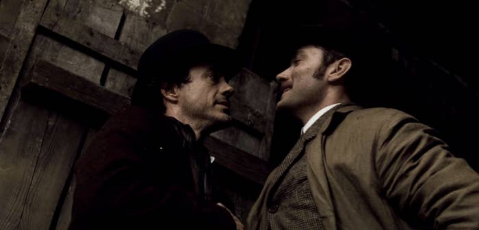 Robert Downey Jr. in a dark coat and hat faces Jude Law in a tan overcoat and hat in a tense scene against a wooden backdrop