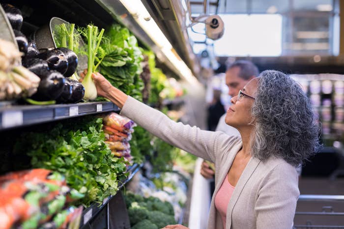 A woman with curly hair and glasses reaches for vegetables in a grocery store aisle. She is wearing a light cardigan over a pink top