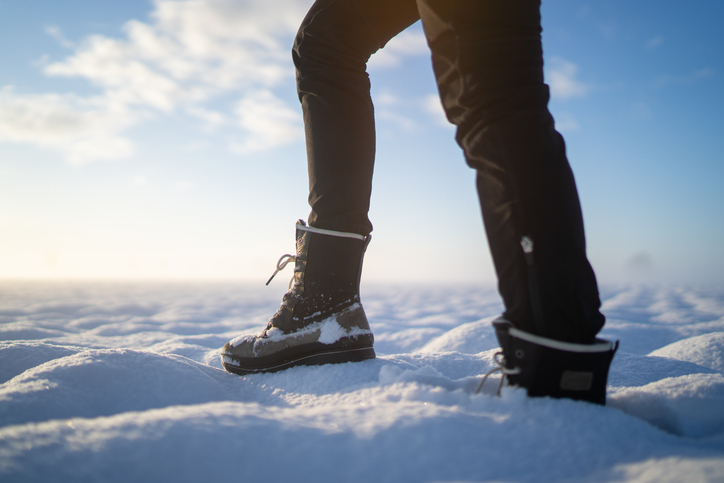 Person walking on snowy terrain with visible snow-covered boots and pants. Sky and clouds in the background