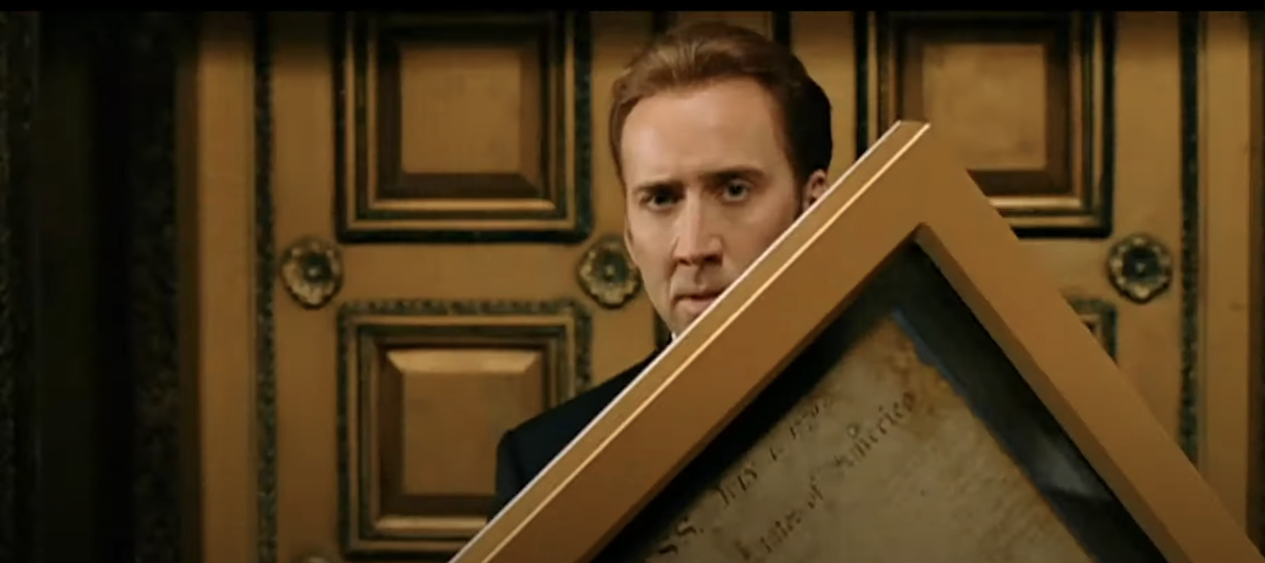 Nicolas Cage is holding a large, framed parchment in front of a detailed wooden door
