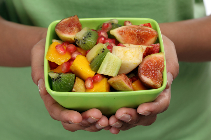 A person holds a green bowl filled with a fresh fruit salad, including figs, kiwi, mango, pomegranate seeds, and other fruits