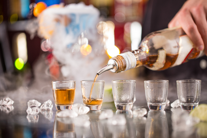 Four shot glasses on a bar being filled with liquor from a bottle, surrounded by ice cubes and some smoke or fog in the background