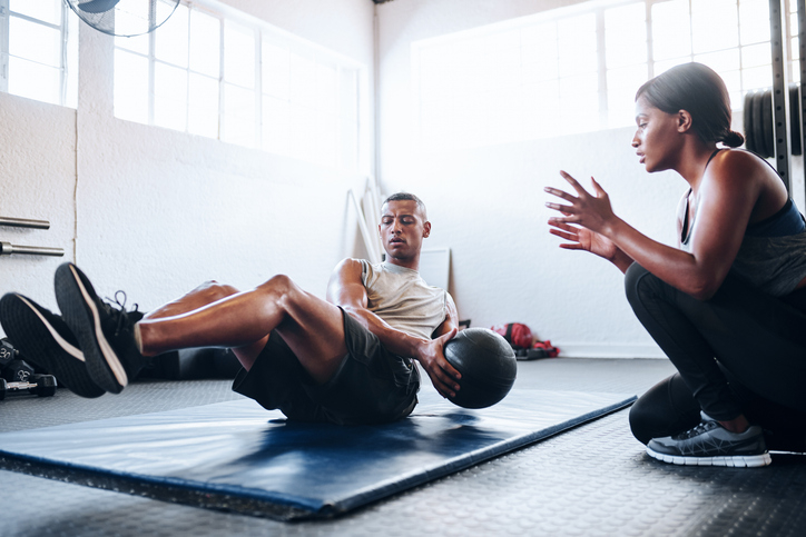 Man holding a medicine ball doing exercises on a mat while a woman kneels beside him, giving instructions in a gym setting