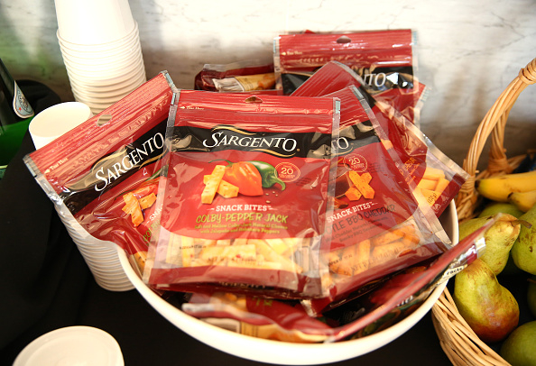 Bowl filled with packages of Sargento Snack Bites Colby-Pepper Jack cheese. Cups and a basket of fruit are seen in the background