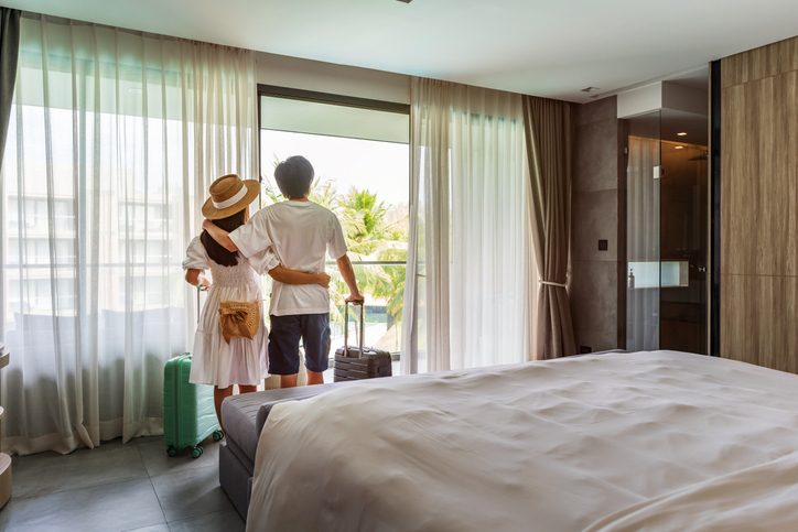 Two people standing in a hotel room, embracing and looking out of a window. One is wearing a hat. They have luggage beside them