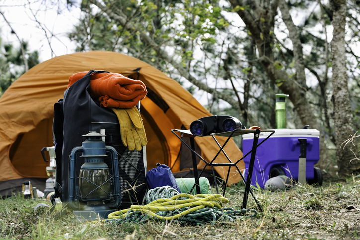 Camping setup with an orange tent, backpack, water jug, cooler, lantern, binoculars, rope, gloves, and a rolled-up sleeping bag in a forested area