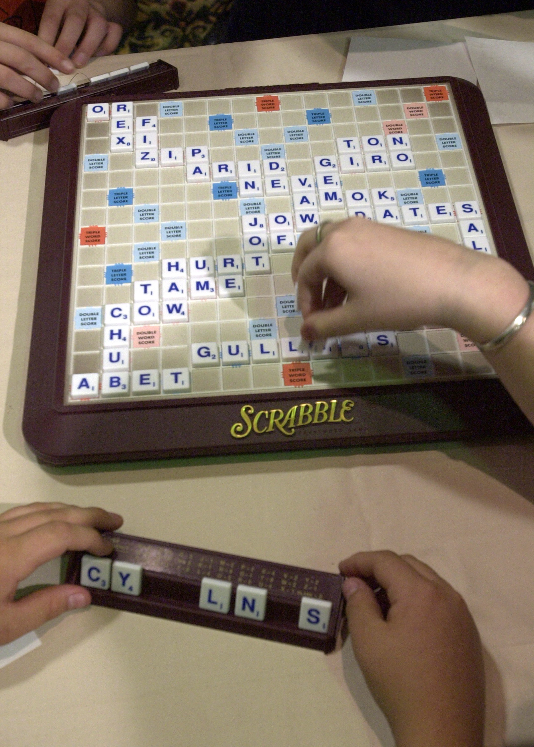 Three hands are placing letter tiles on a Scrabble board, with words like "Gullies" and "Pirated." Another hand is holding a rack with tiles spelling "CYLNS."