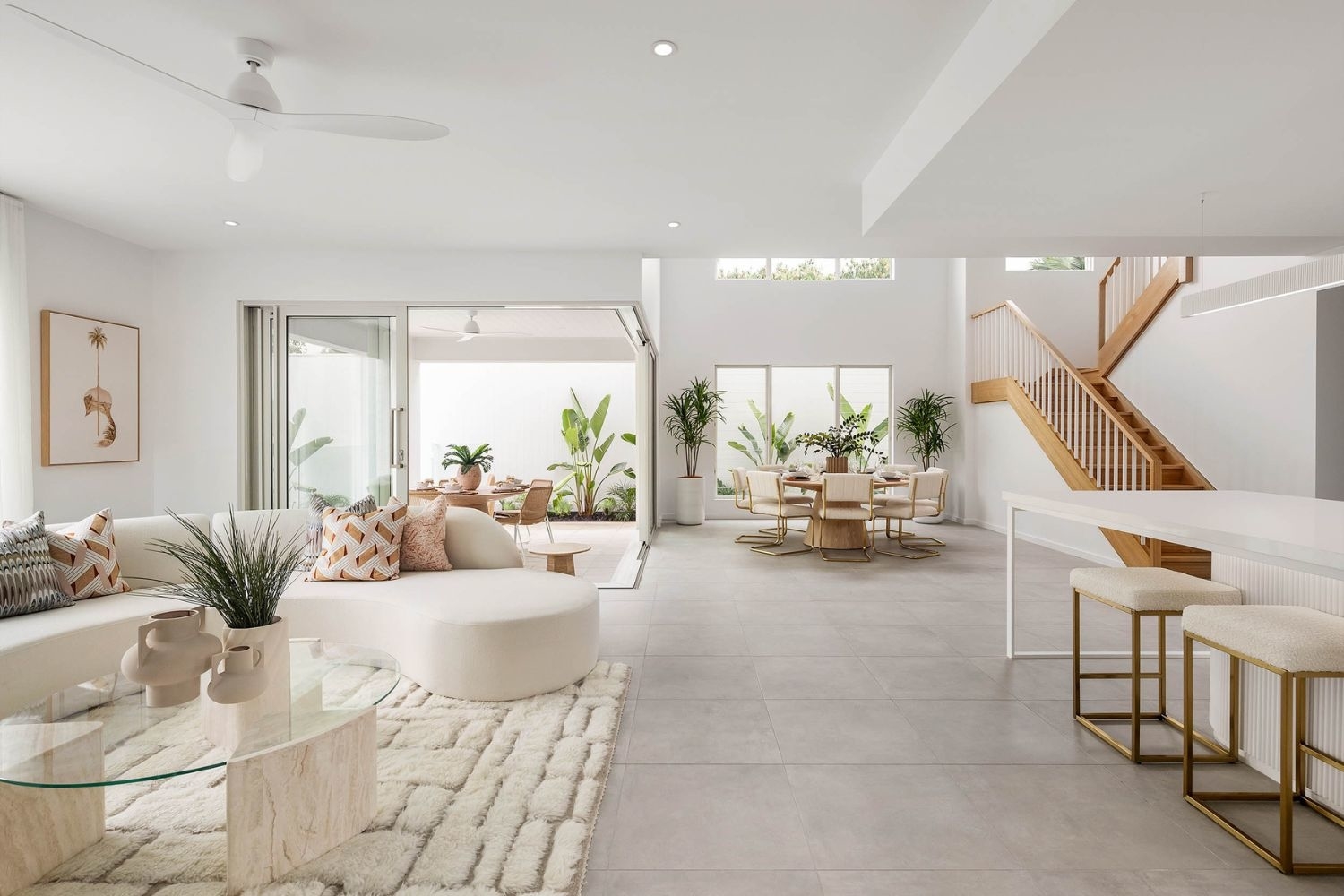 Modern open-plan living and dining area with minimalist furniture, a glass coffee table, and decorative plants. A staircase leads to an upper level