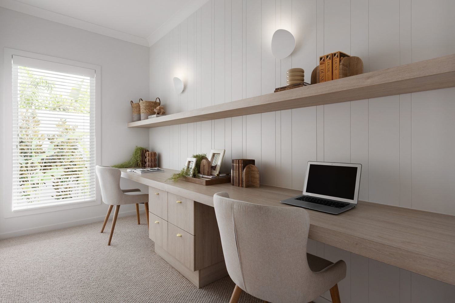 A modern home office with two workstations, each with a chair, under a long shelf with various baskets and decor items. A window with blinds is on the left