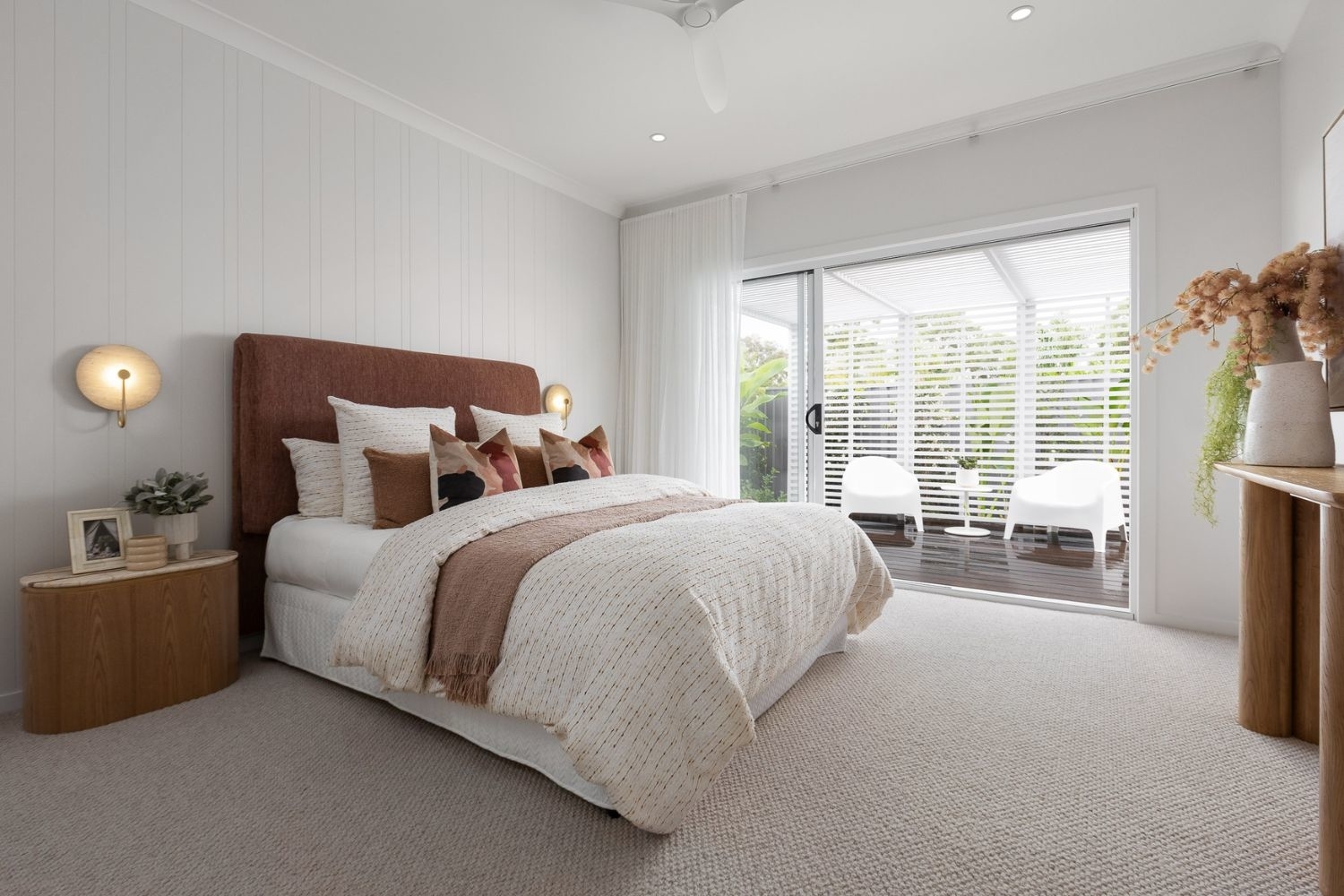 A modern bedroom with a neatly made bed, cushions, a bedside table with decor, soft lighting, and a sliding door opening to a patio with lounge chairs