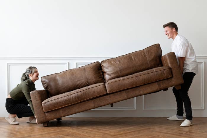 A woman and a man are smiling and lifting a brown leather couch in a room with wooden flooring and a white wall