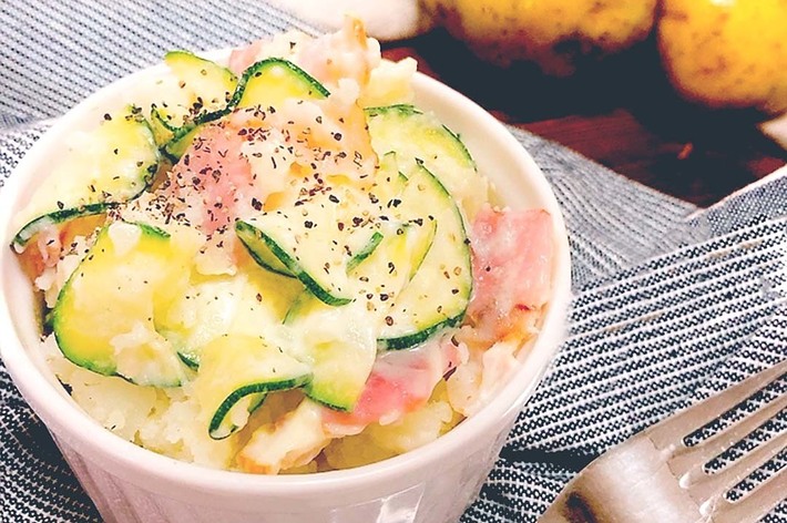白いカップに入ったジャガイモときゅうりのポテトサラダが置かれた写真です。カップの隣にフォークがあります。背景にはジャガイモが写っています。