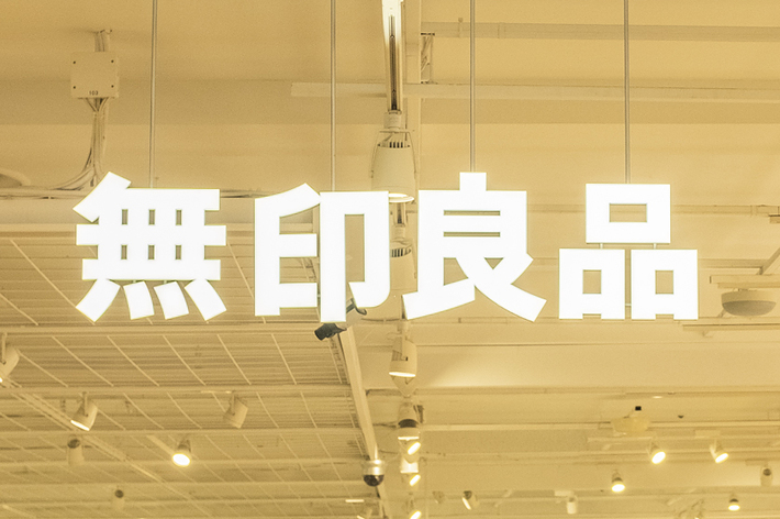 Interior view of a Muji store with people browsing shelves filled with various products. Japanese signage hangs from the ceiling