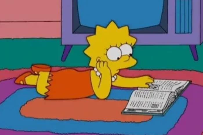 Lisa Simpson lies on a rug, reading a book, with a television in the background