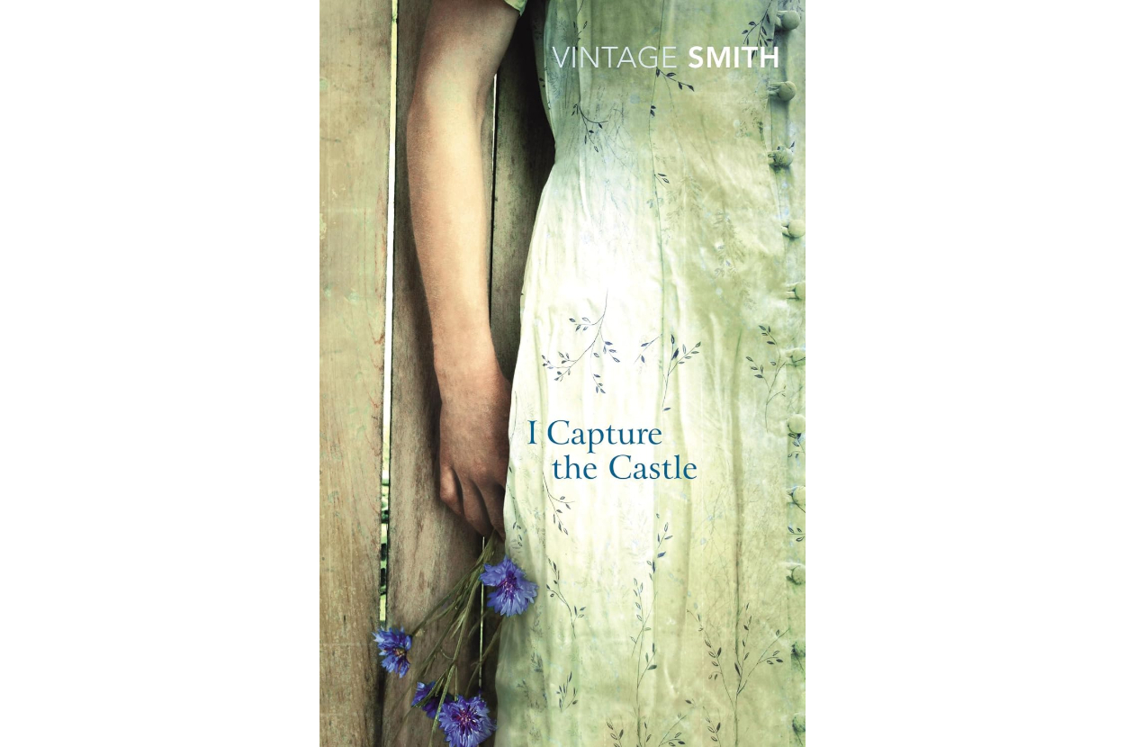 Book cover of &quot;I Capture the Castle&quot; by Dodie Smith, showing part of a woman's arm and hand holding blue flowers, next to a rustic wooden door