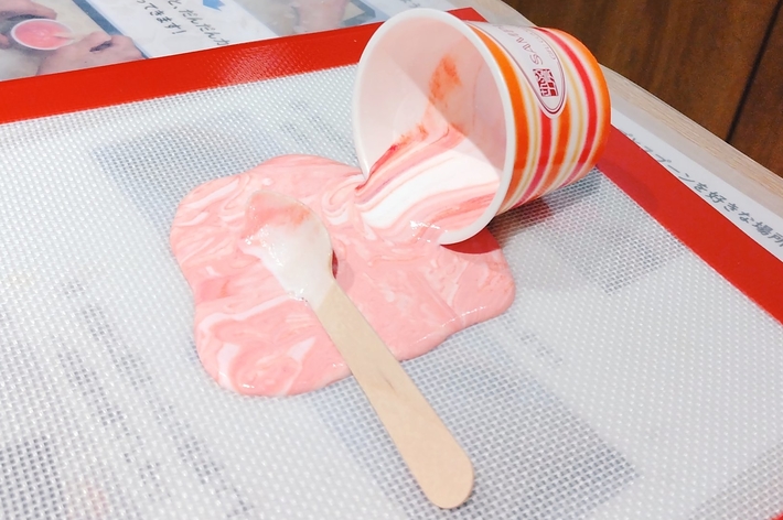 倒れたカップからピンク色のアイスクリームが溶け出し、アイスの棒が手前に置かれている。