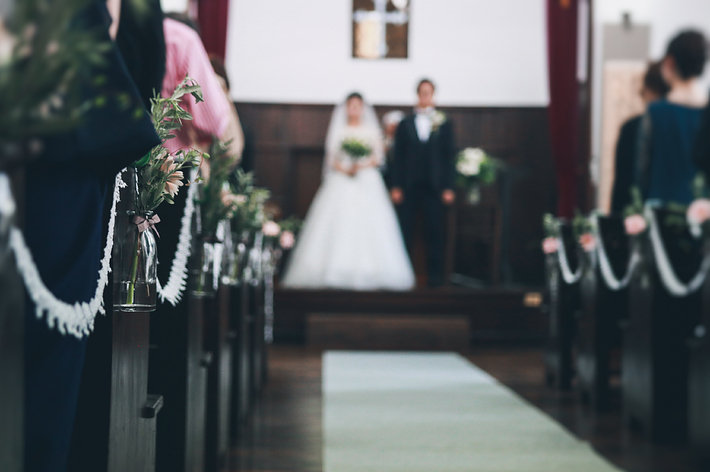 新郎新婦が結婚式で祭壇に立っている。両側の椅子には花が飾られている。新婦はドレス、新郎はスーツを着ている。