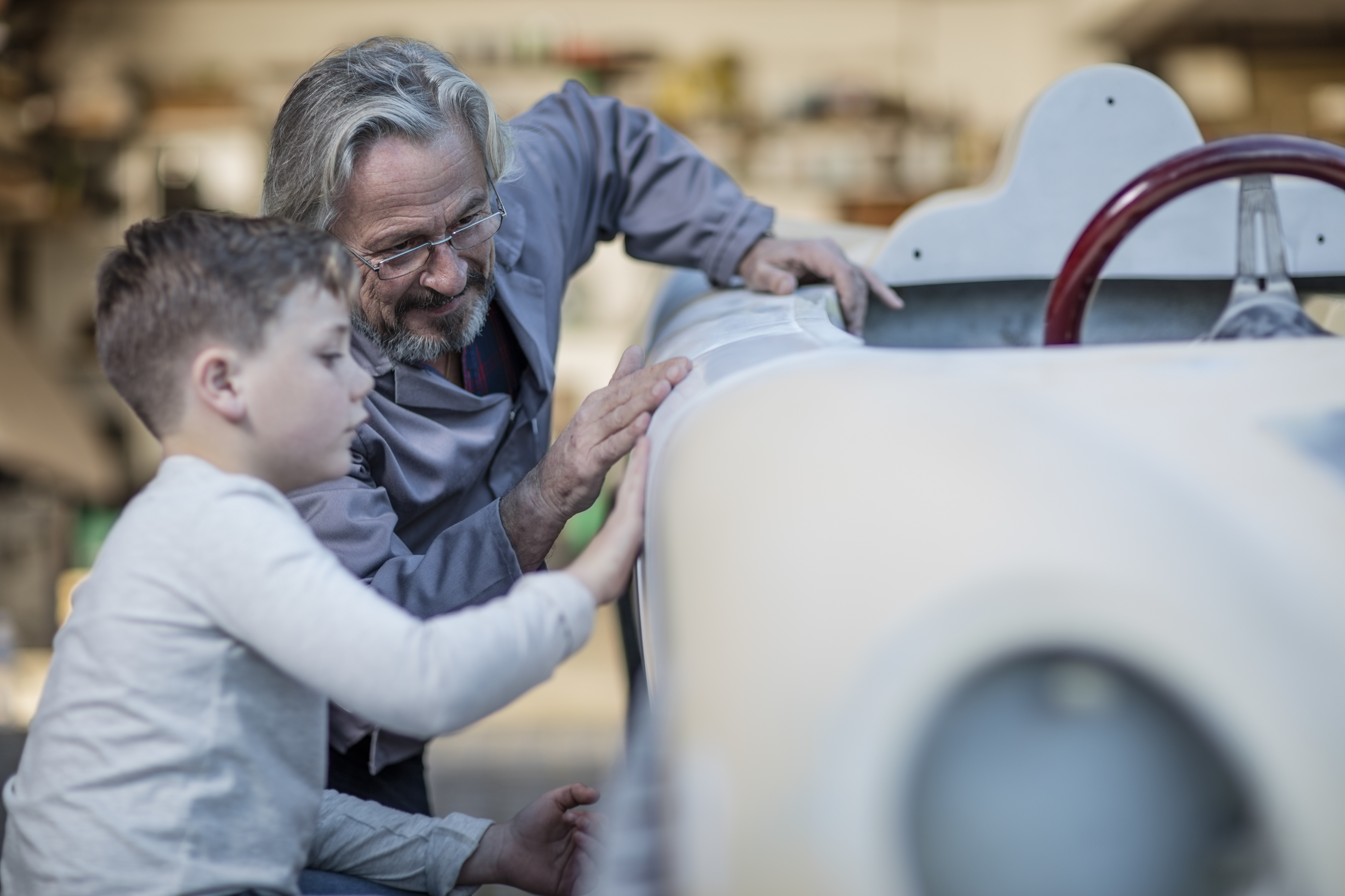 An older man with gray hair and a beard, wearing glasses, and a young boy examine the surface of a vintage car together in a garage