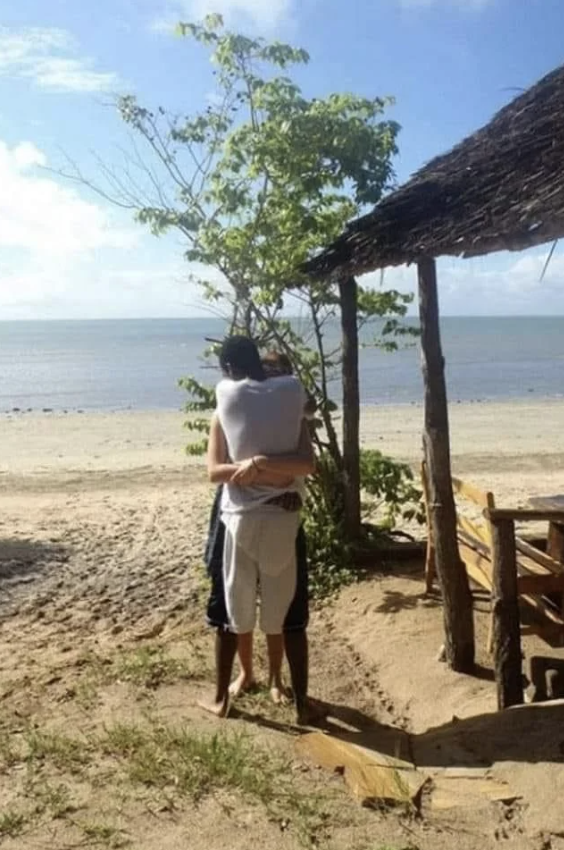 Two people in a warm embrace near a thatched hut on a beach with a tree and the ocean in the background