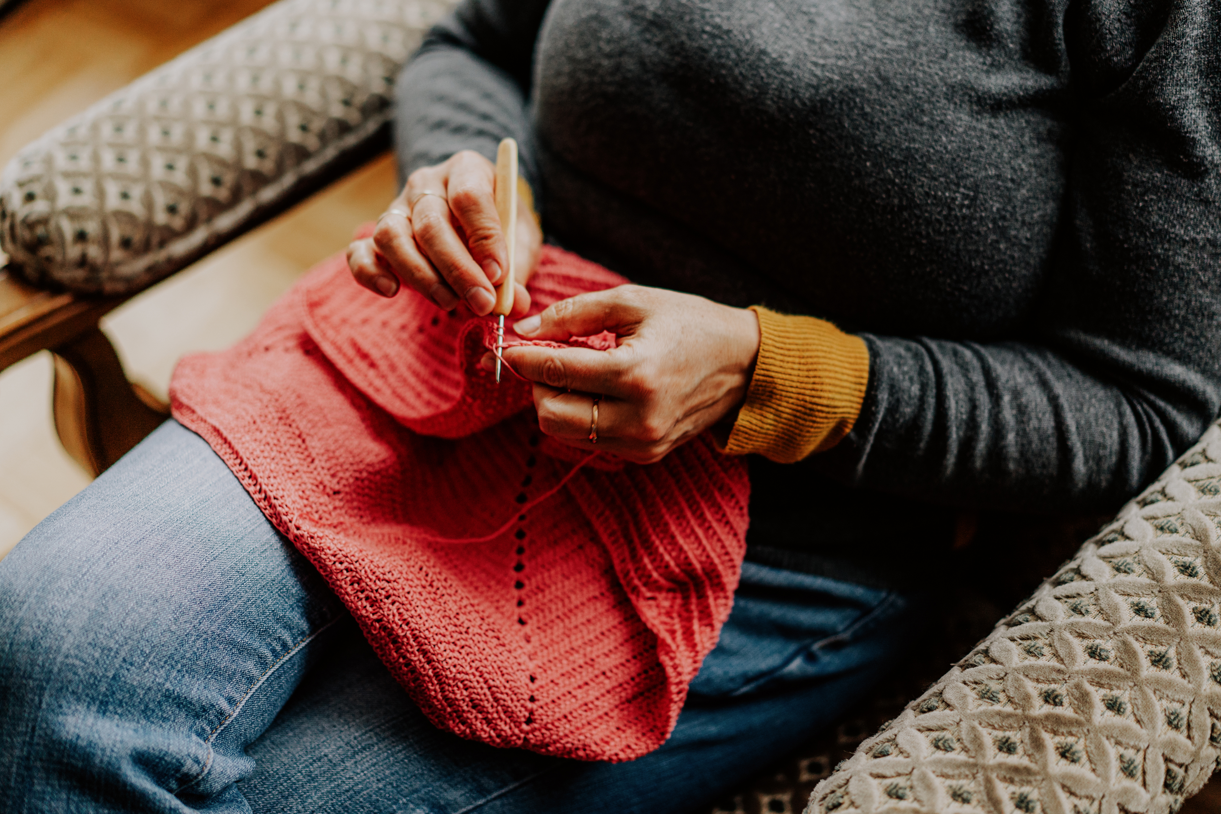 A person sits in a patterned chair, knitting with red yarn on their lap. Only their hands and torso are visible, wearing a gray sweater and jeans