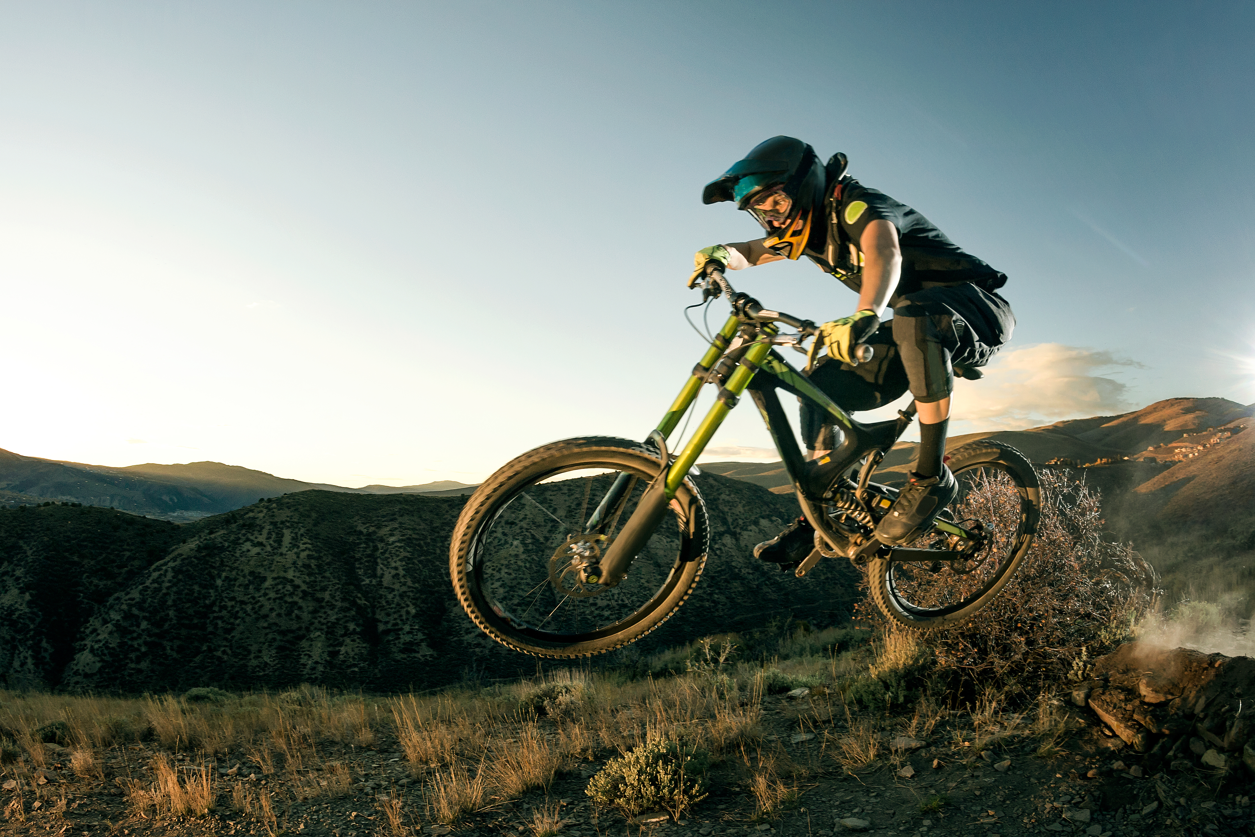 A person wearing protective gear is mid-air while performing a jump on a mountain bike in a rugged, mountainous landscape