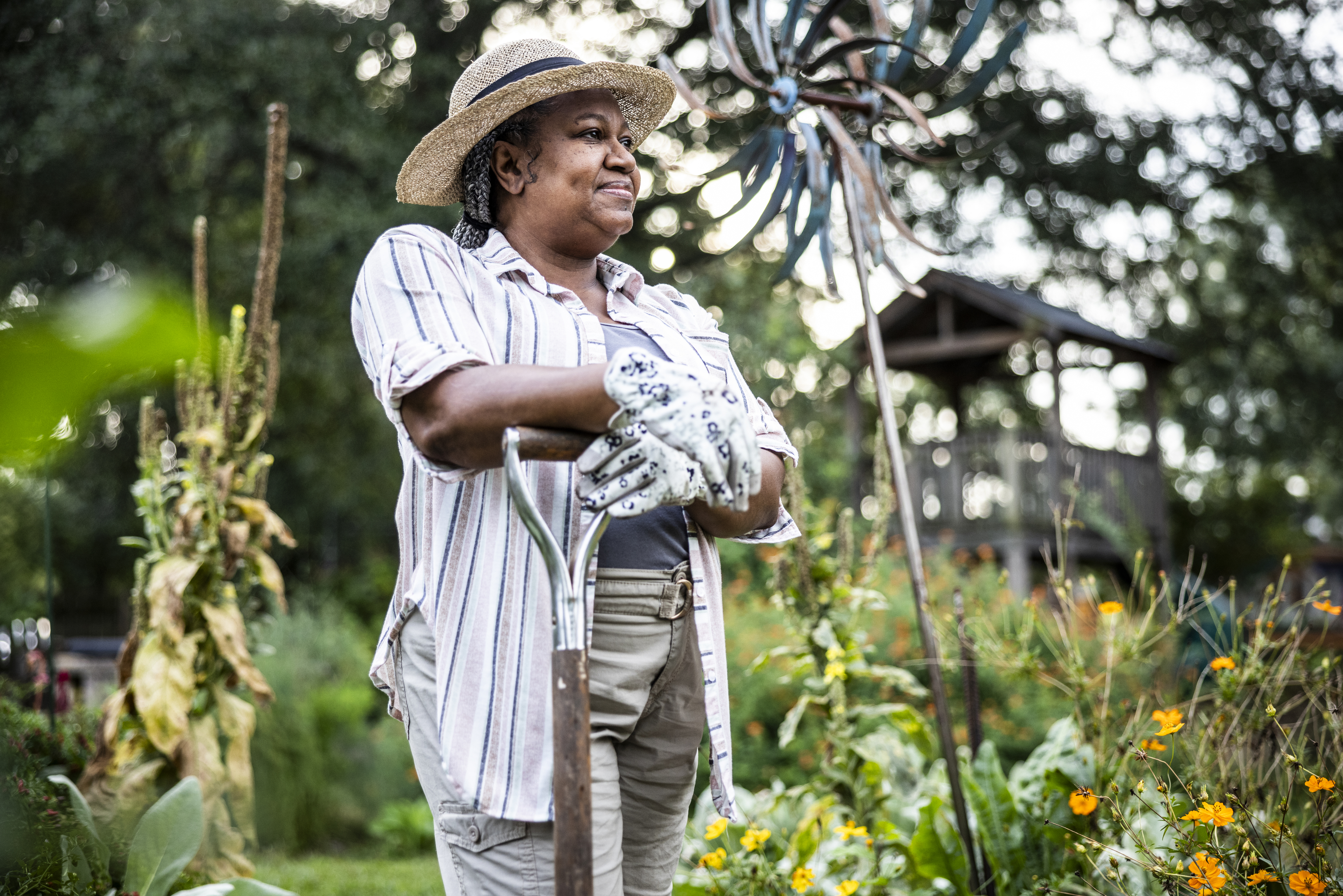 A woman dressed in a striped shirt, khaki pants, gloves, and a straw hat stands in a garden, holding a shovel, with plants and flowers around her
