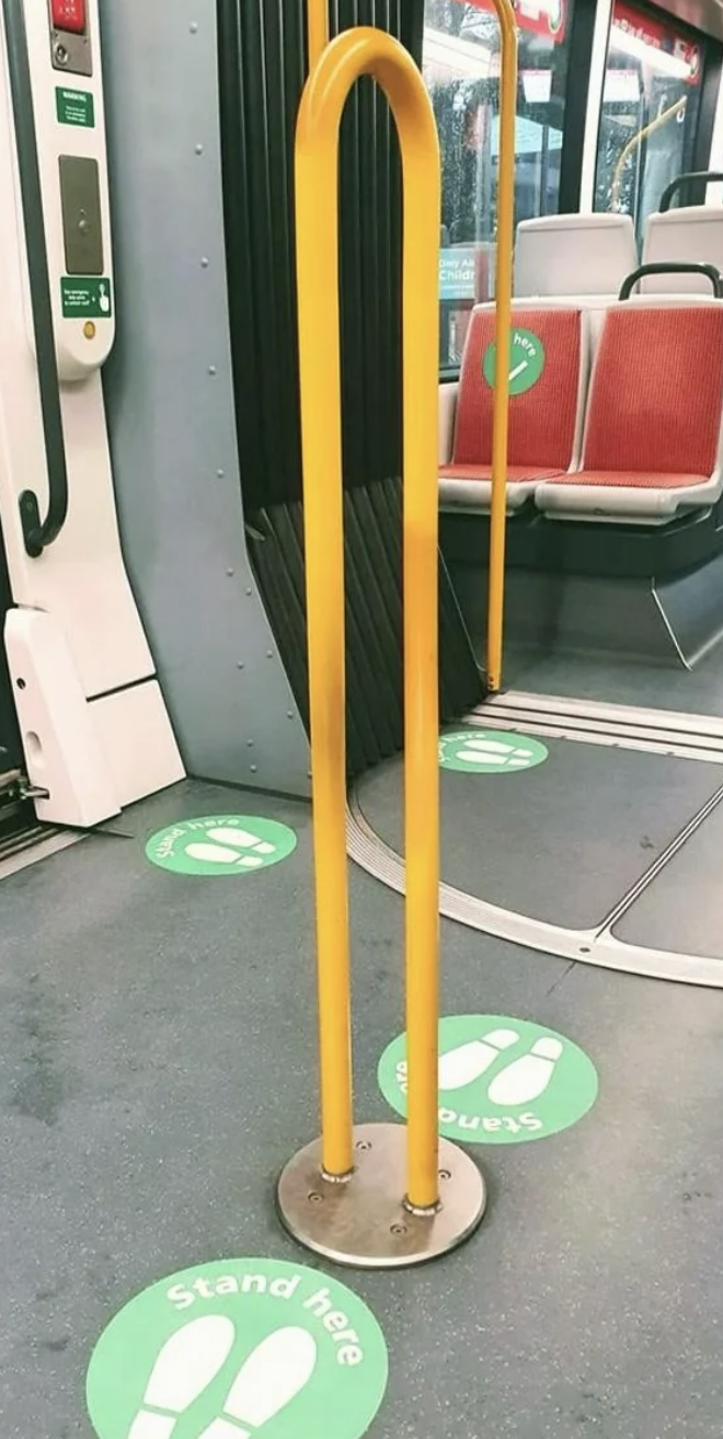 Interior of a bus with "Stand Here" markings on the floor and a yellow handrail for standing passengers