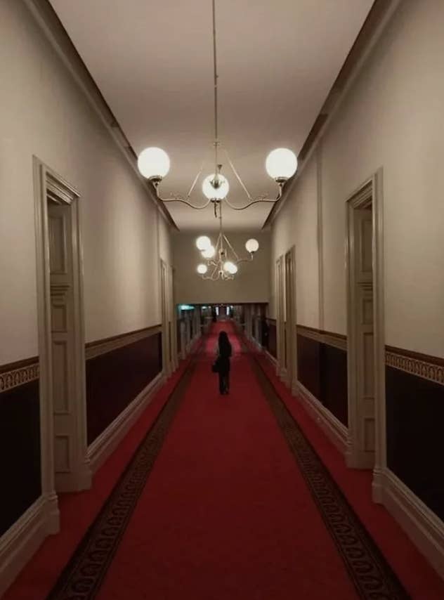 Person walking down a long, dimly lit corridor with ornate light fixtures, red carpet, and high ceilings. The walls have decorative molding and dark panels