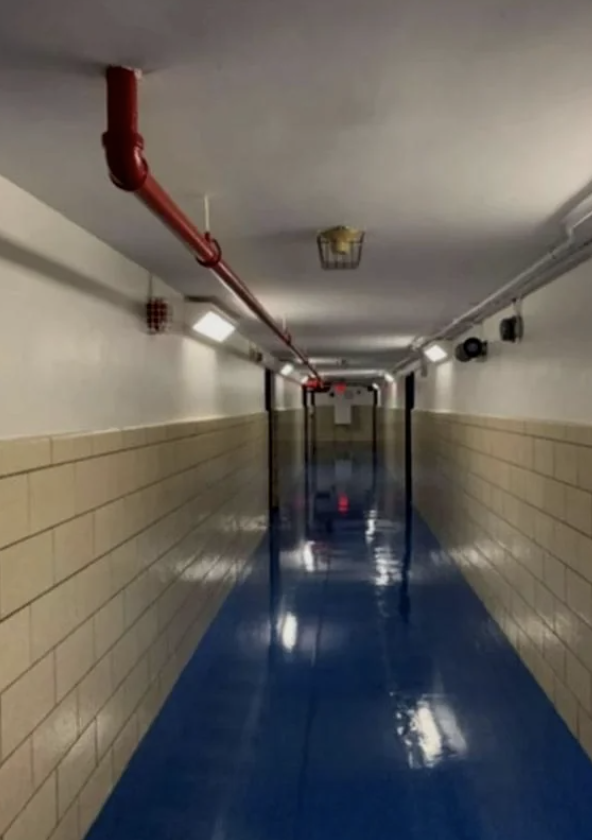Empty, well-lit, long hallway with blue floors, beige walls, and visible pipes on the ceiling. No persons or text are present in the image