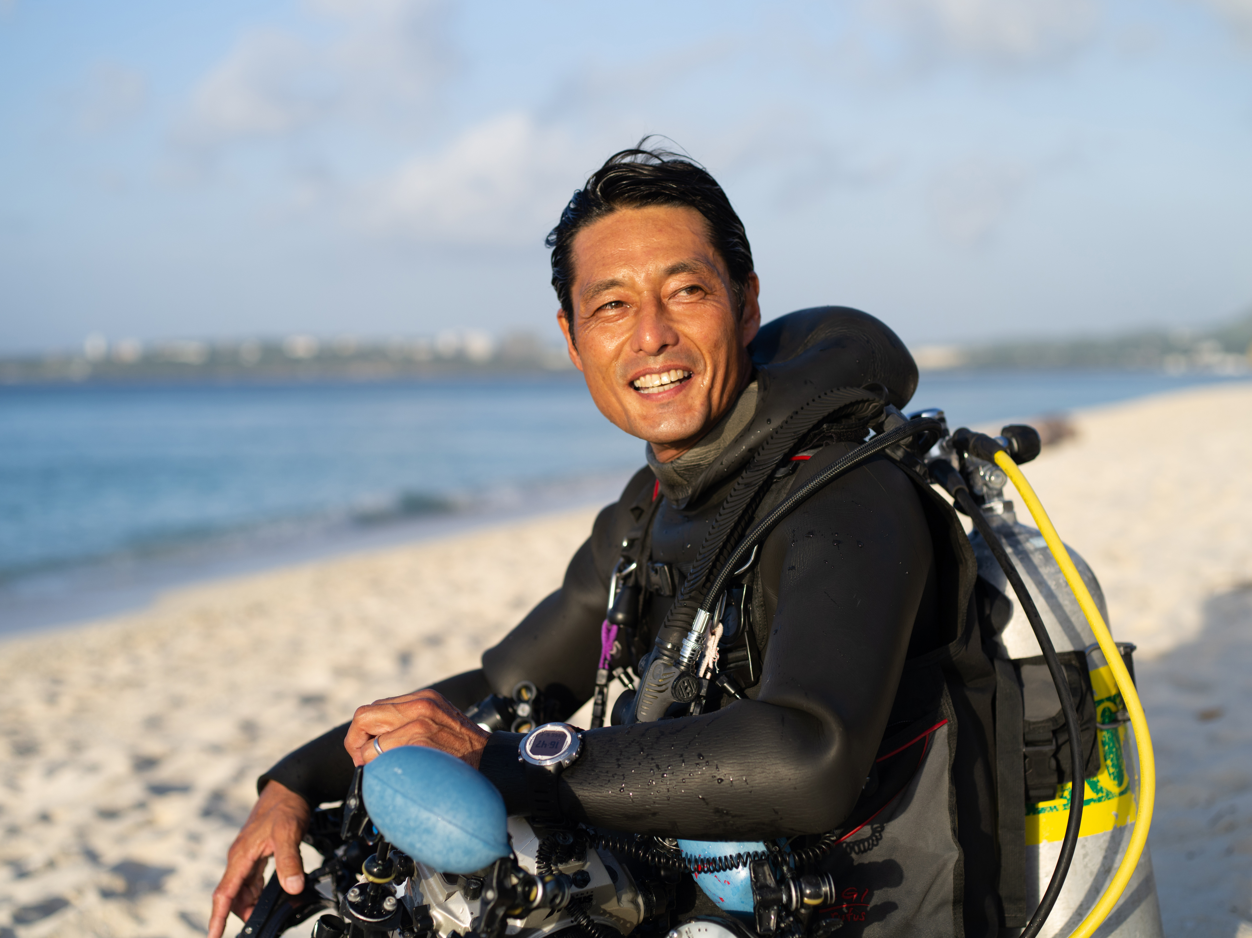 A person sits on a sandy beach in scuba gear, smiling and holding diving equipment. The ocean and sky are visible in the background