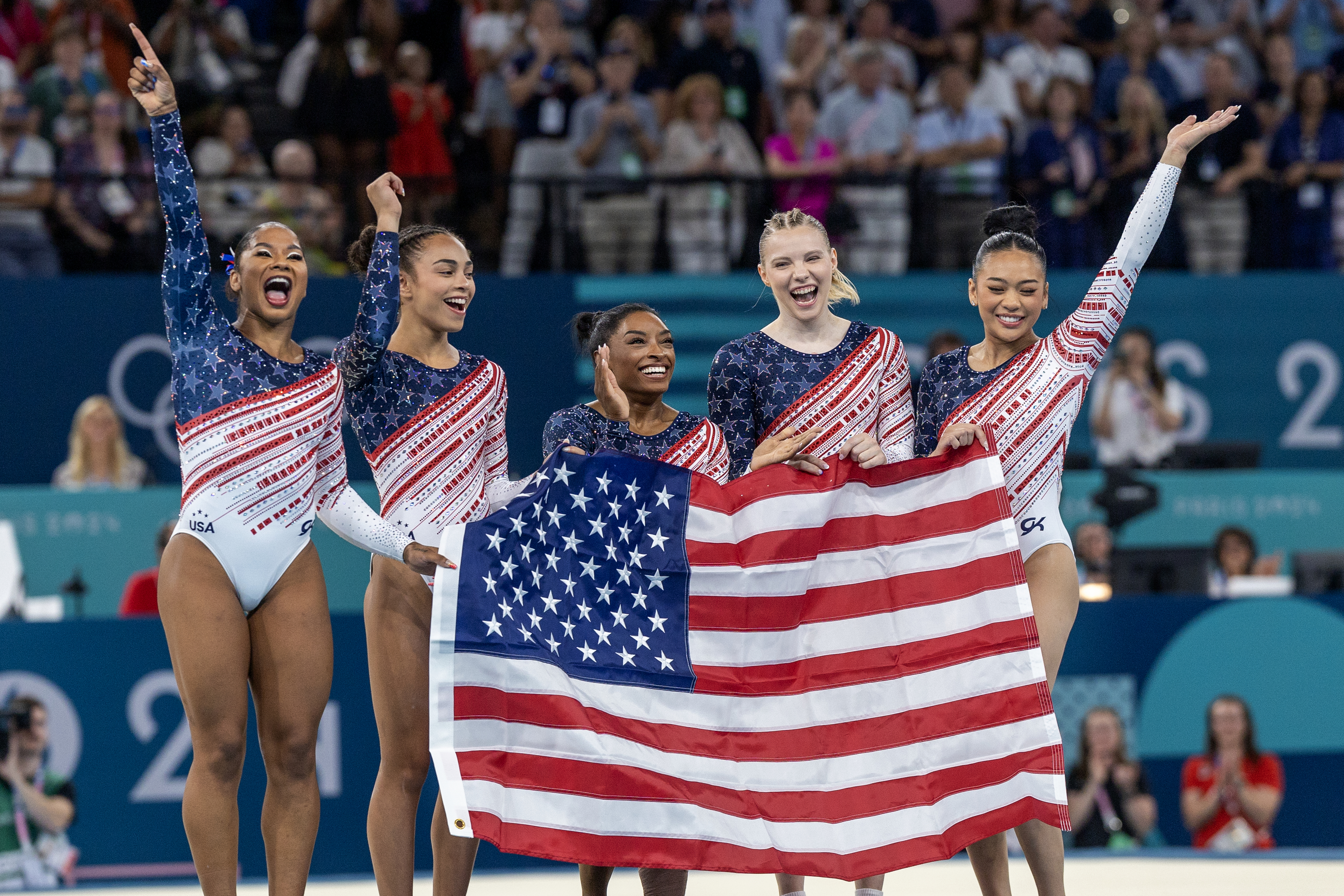 Simone Biles, Jordan Chiles, Sunisa Lee, Grace McCallum, and Mykayla Skinner stand together, smiling and holding an American flag after a gymnastics competition