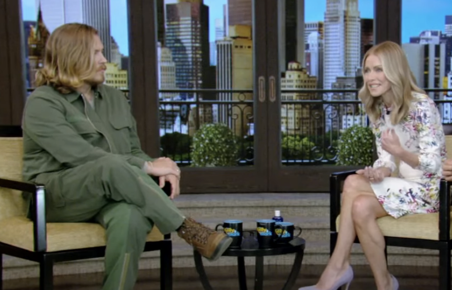 Dylan Sprouse in a green suit and Kelly Ripa in a floral dress on a talk show set, engaged in conversation