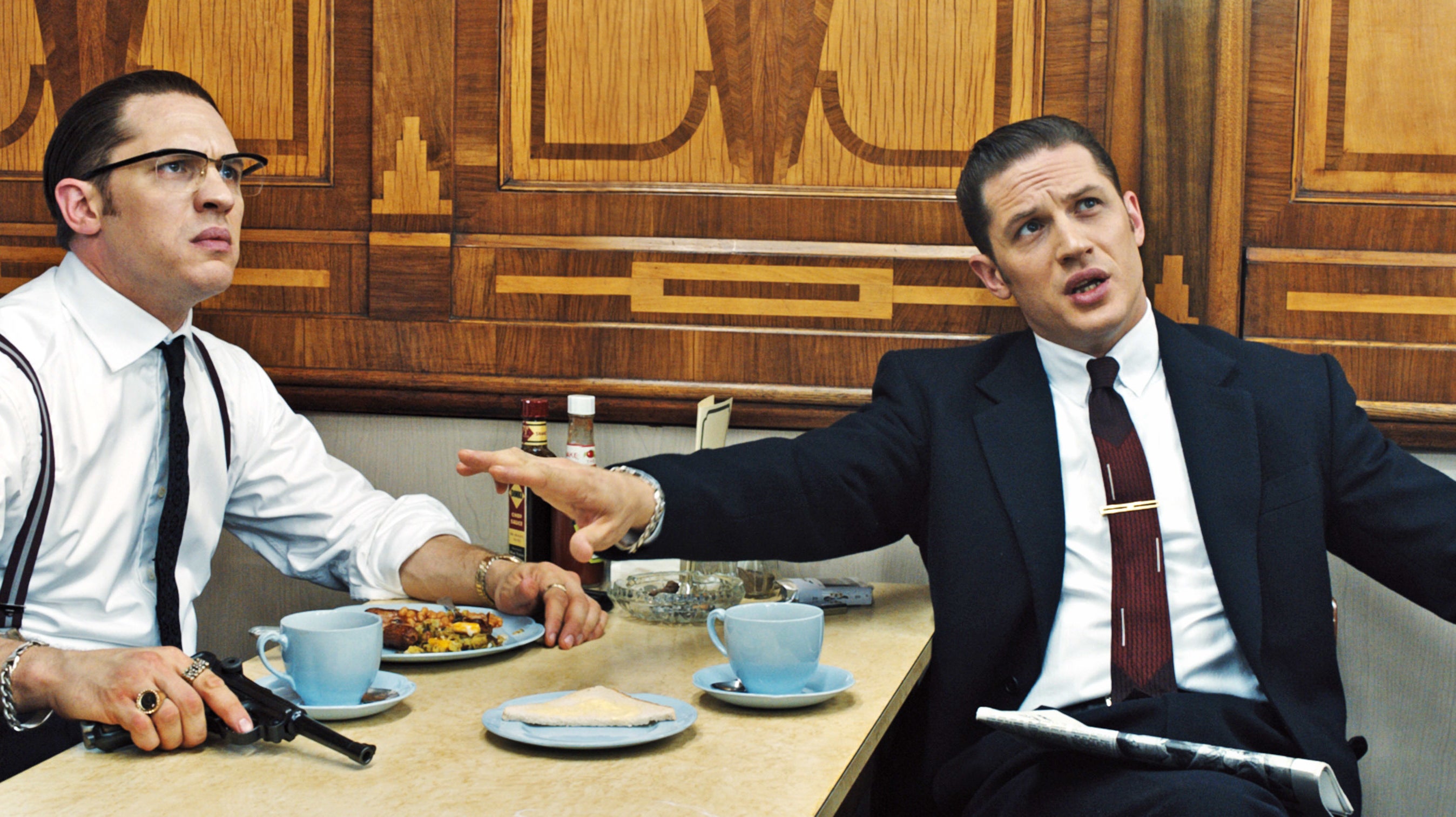 Tom Hardy, dressed in a suit and tie with glasses, and Tom Hardy, dressed in a suit and tie, sit at a table with food and drinks. One has a gun out