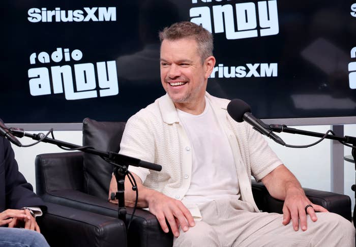 Matt Damon, in a white shirt and pants, smiles while sitting for an interview on SiriusXM's Radio Andy