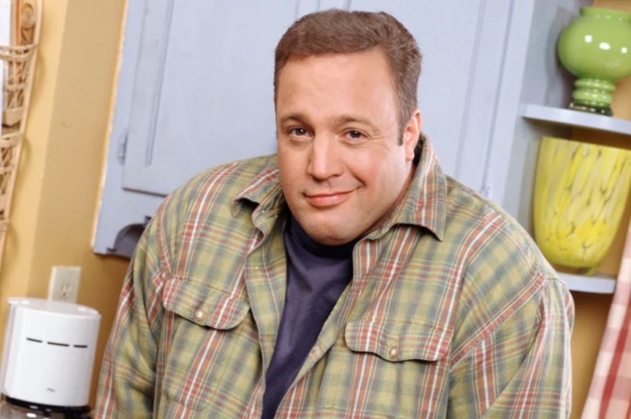 A man wearing a plaid shirt and a dark undershirt stands in a kitchen with a slight smile, posing near a shelf with decorative items