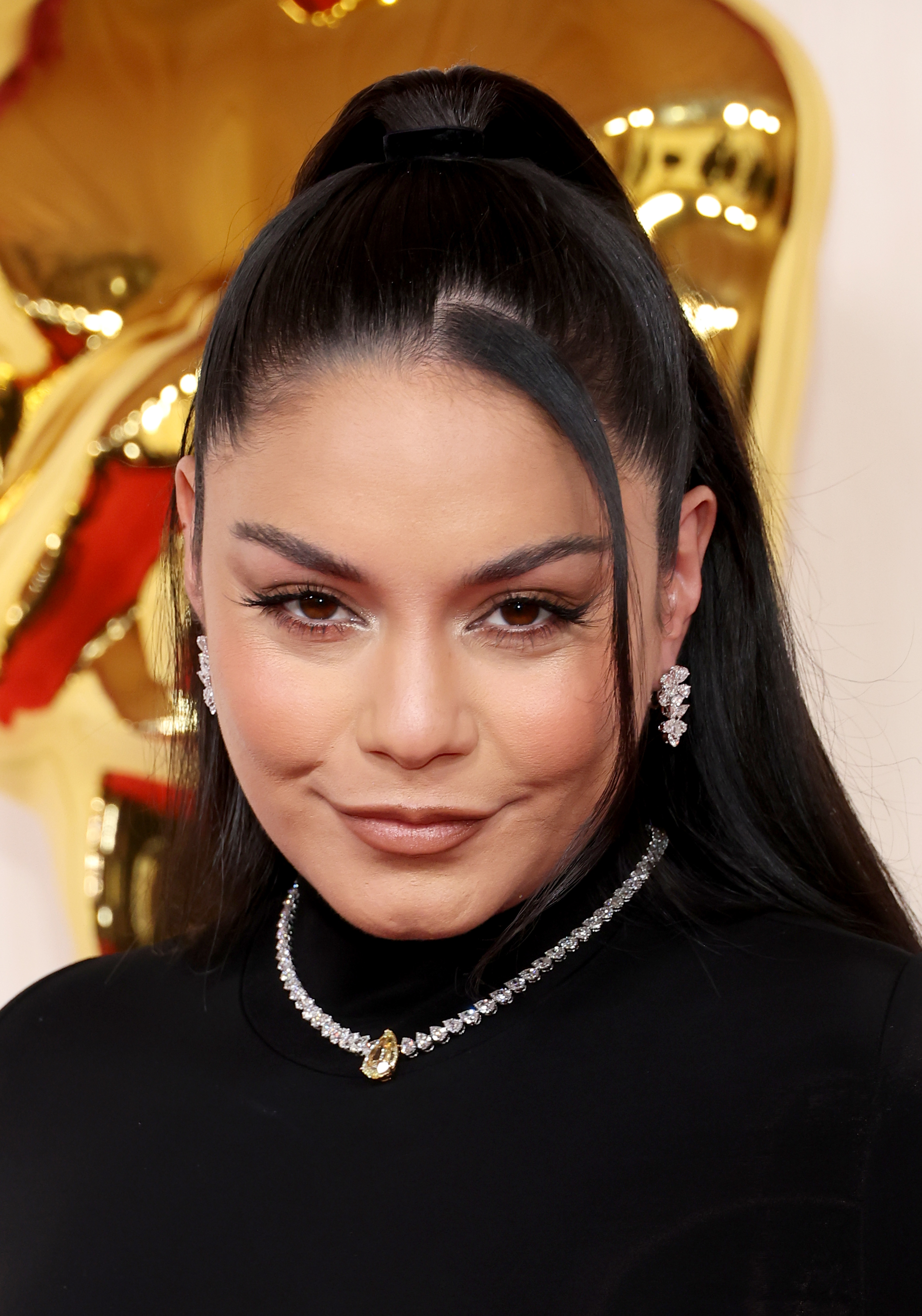 Vanessa Hudgens on the red carpet, wearing a sleek black outfit accessorized with diamond earrings and a necklace. Her hair is styled in a high ponytail