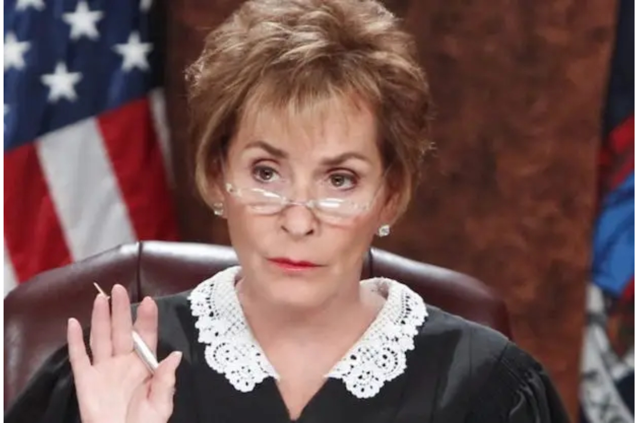Judge Judy Sheindlin in judicial robes, holding a pen, and raising her right hand, in front of a U.S. flag