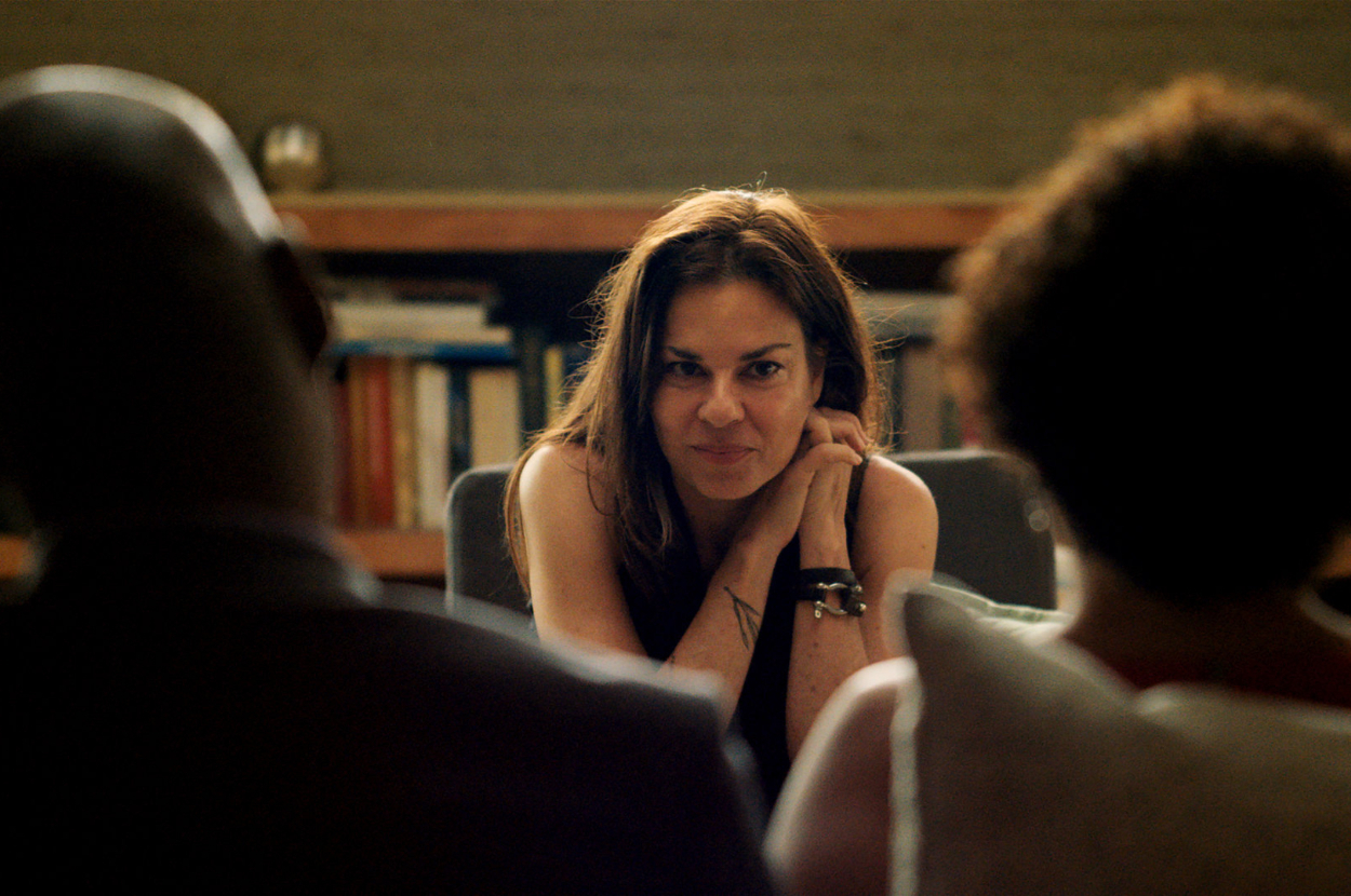 Three people sitting in a living room, with a focused woman in the center. The other two people's backs are to the camera