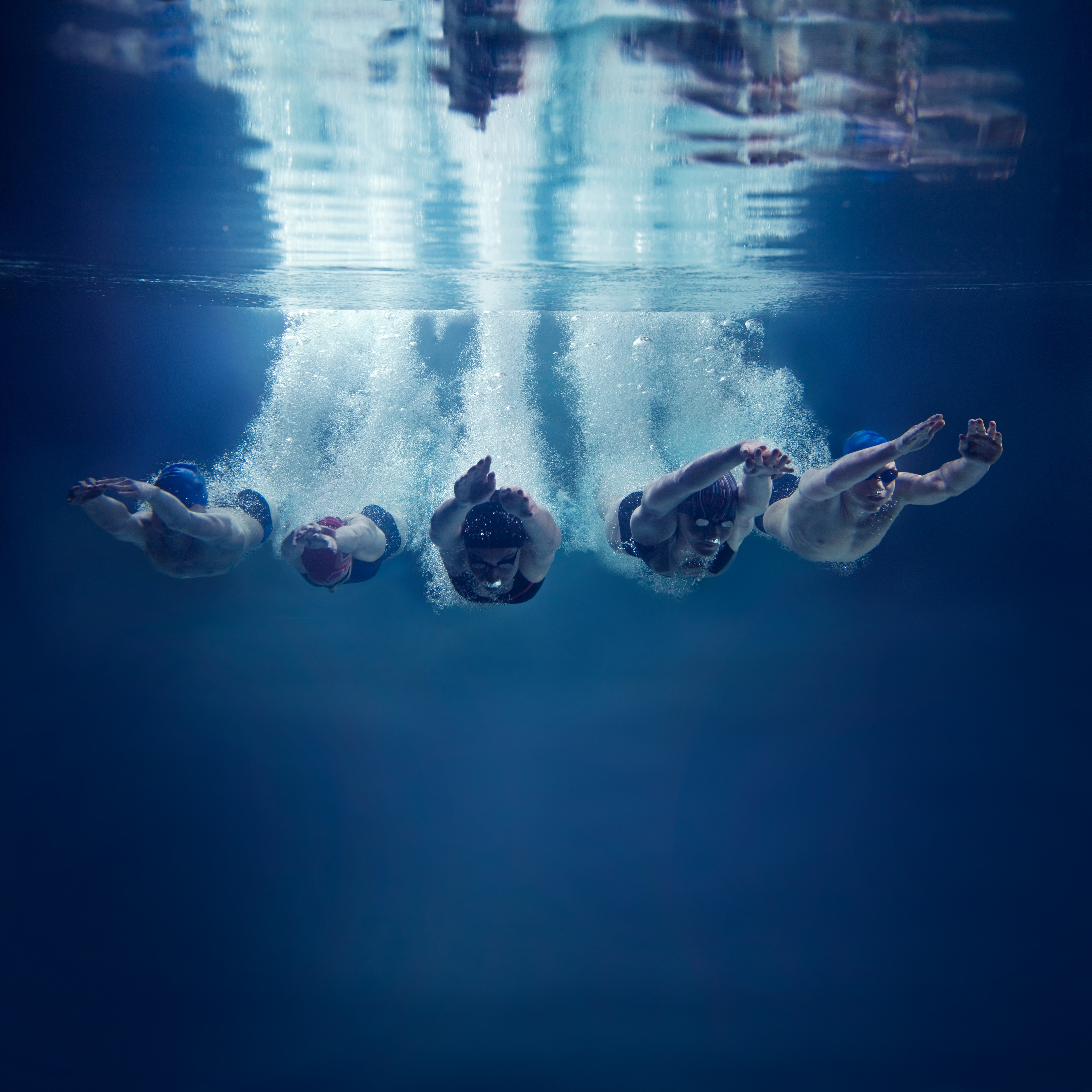 Five swimmers diving into a pool in unison, creating splashes as they enter the water. The image captures them from an underwater perspective