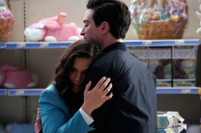 America Ferrera in a blue blazer embraces Ben Feldman in a store aisle filled with plush toys and gift baskets