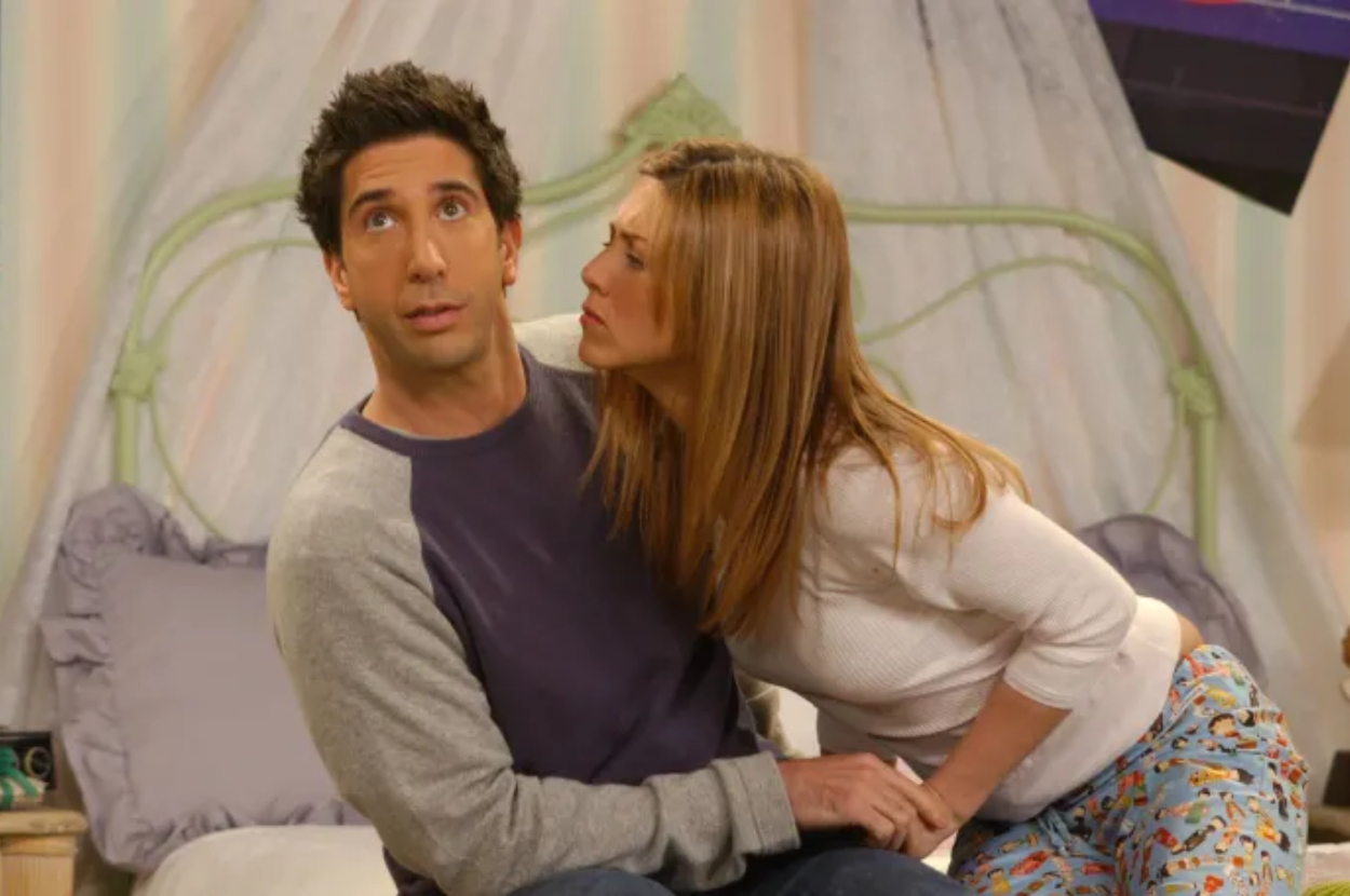 David Schwimmer making a funny expression while Jennifer Aniston looks at him with concern in a bedroom scene from the TV show "Friends."