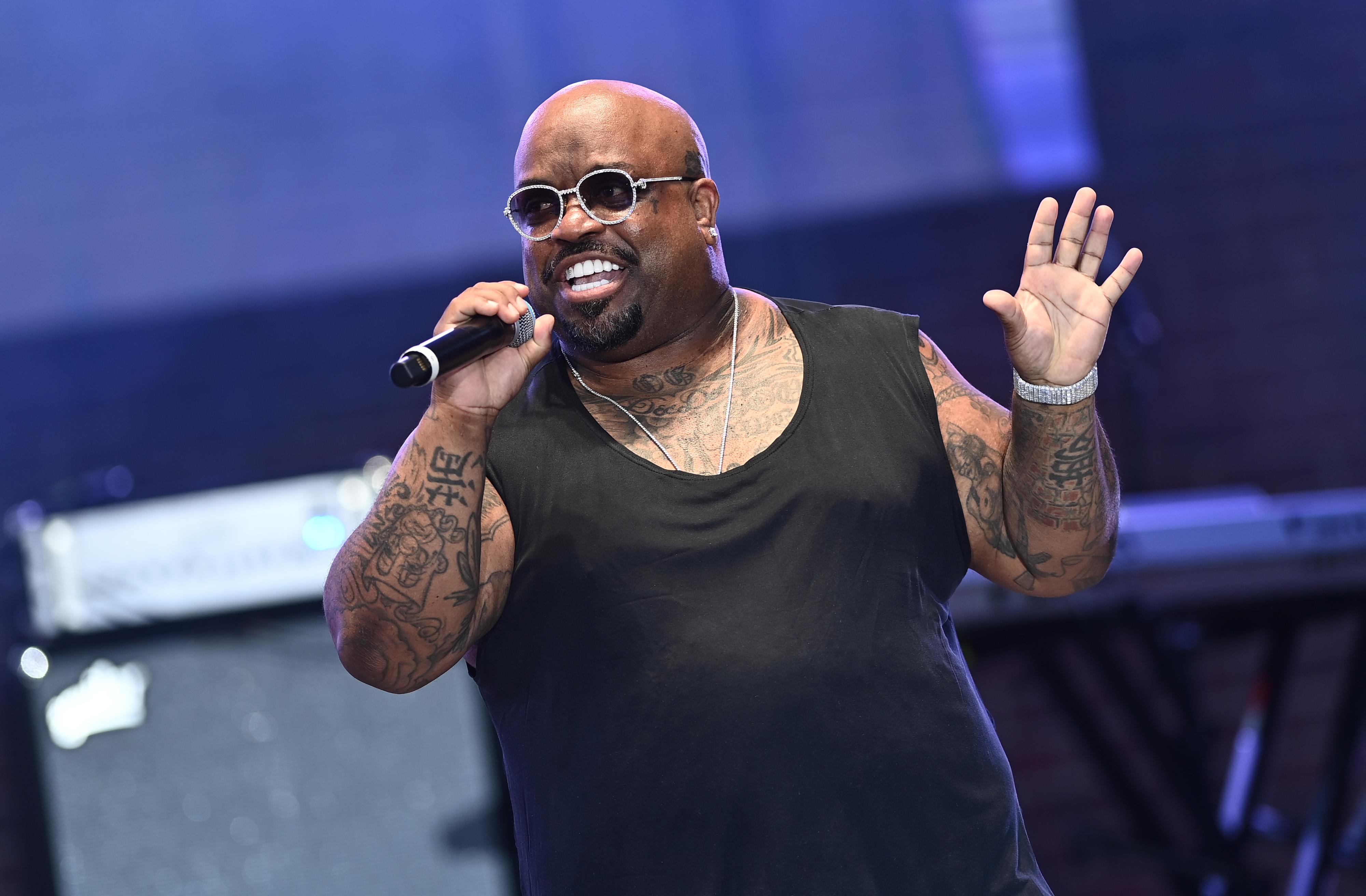 Cee-Lo Green singing onstage holds a microphone and waves to the audience. He wears a sleeveless shirt, sunglasses, and has tattoos on his arms