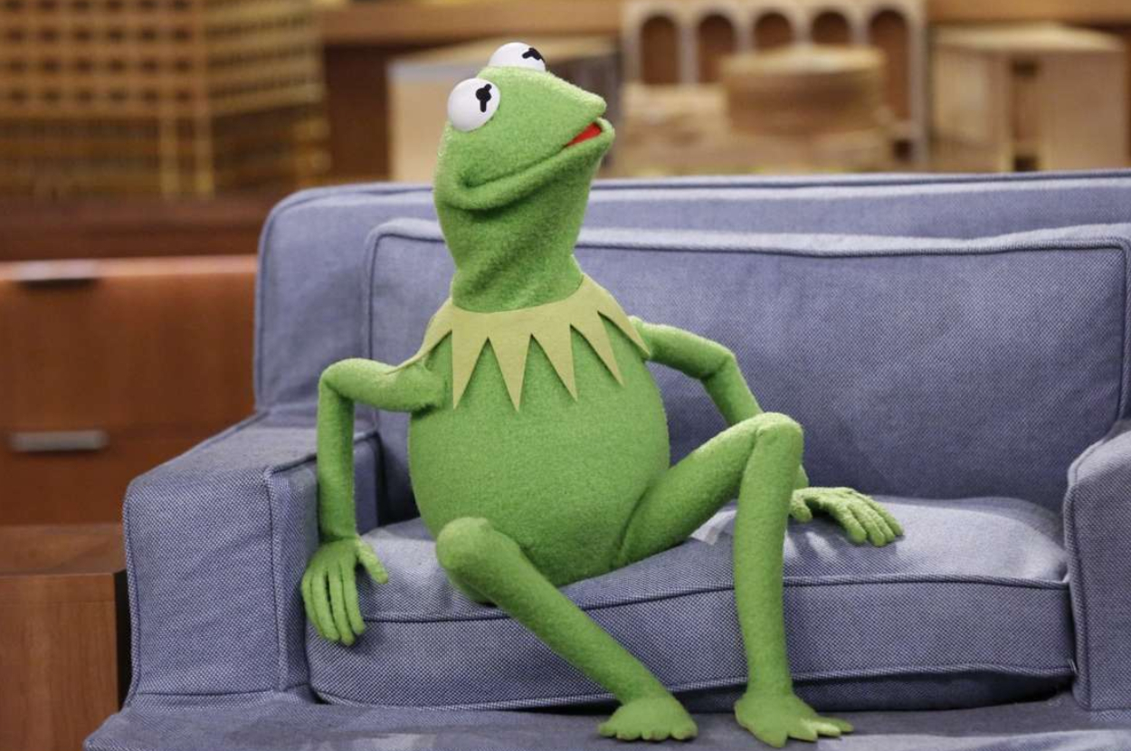 Kermit the Frog sitting on a blue couch, looking thoughtful with one arm resting on the couch's back and the other on the seat