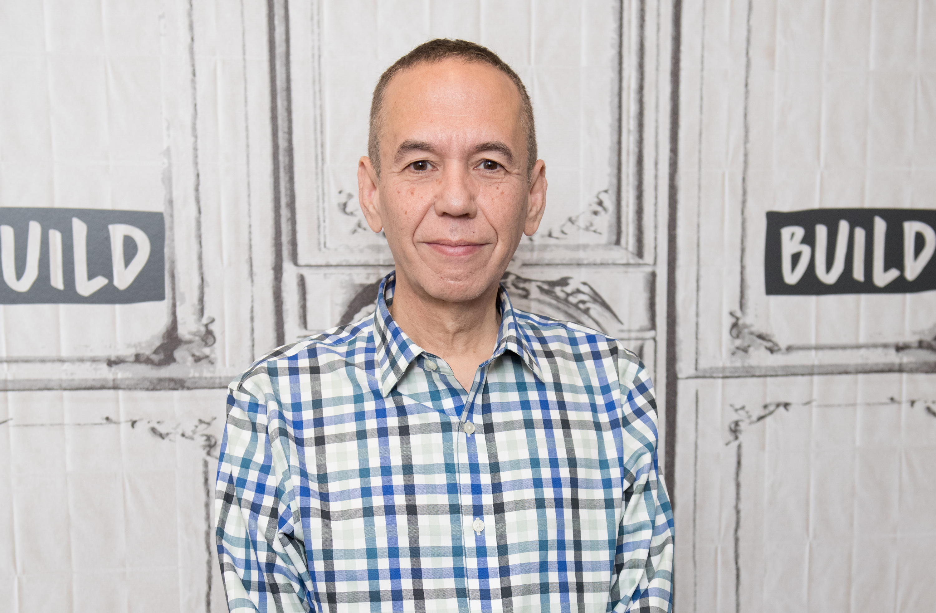Gilbert Gottfried, smiling, with short hair, wearing a checkered shirt, stands in front of a backdrop with the word "BUILD" on it