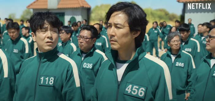 A crowd of people wearing green tracksuits with numbers on them, including 118, 456, and 246. The scene appears to be from the Netflix series "Squid Game."