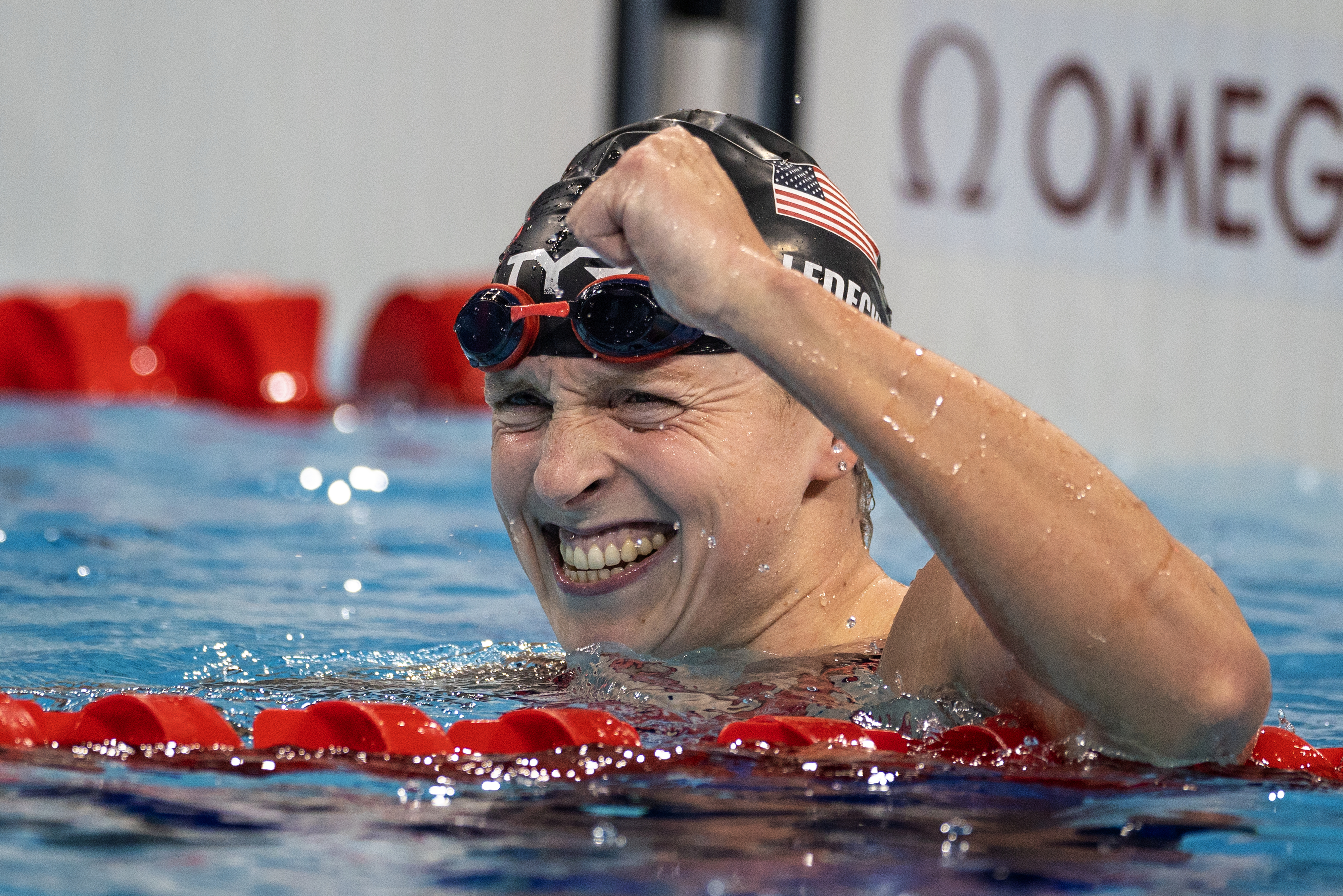 Katie Ledecky, wearing a swim cap and goggles, celebrates in a pool after a race with a triumphant fist pump and a big smile