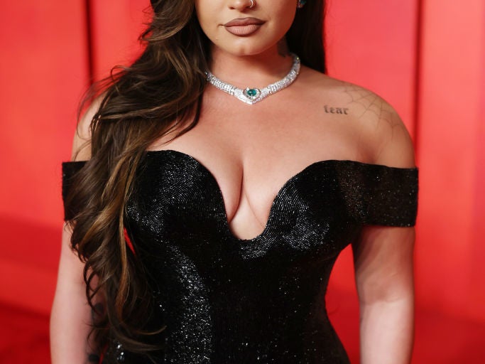 Demi Lovato on a red carpet in an off-the-shoulder, fitted black gown with a sweetheart neckline, accessorized with a diamond necklace