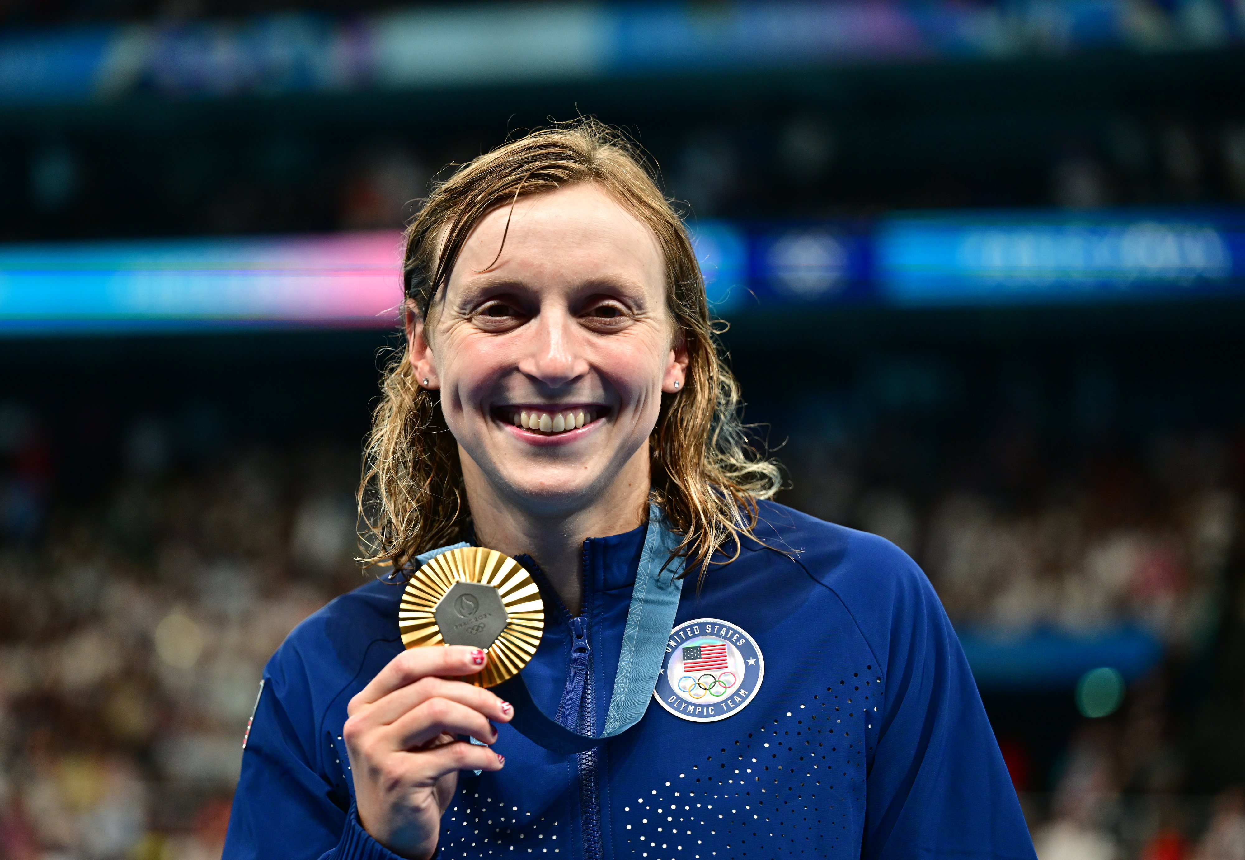 Katie Ledecky smiles, holding her Olympic gold medal, wearing a Team USA jacket, in a crowded stadium