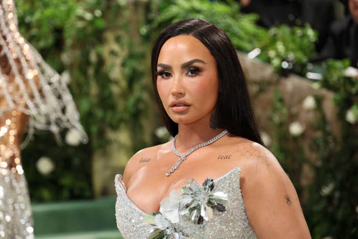 Demi Lovato on the red carpet in a sparkling, form-fitting gown adorned with floral embellishments on the shoulders and chest, with visible tattoos on her collarbone