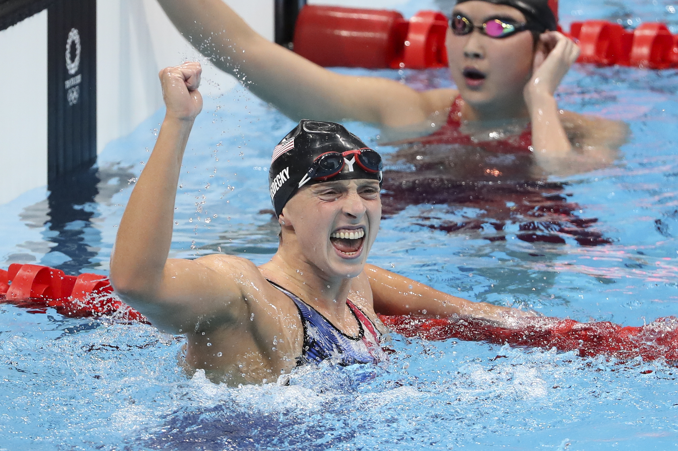 Swimmer Katie Ledecky, with goggles on her head, celebrates in the pool, raising her fist, while another swimmer, likely her competitor, is in the background